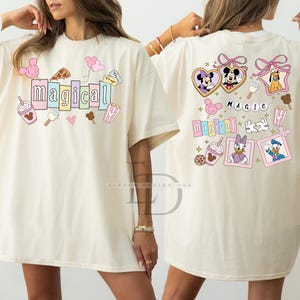 Op de afbeelding: Cr&egrave;mekleurig T-shirt met het woord "magical" in pastelkleuren, omringd door cartoonafbeeldingen van eten en personages. De achterkant van het shirt toont meer cartoonpersonages en het woord "magic".