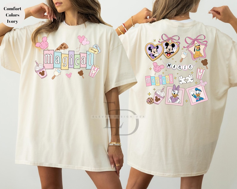 Op de afbeelding: Cr&egrave;mekleurig T-shirt met het woord "magical" in pastelkleuren, omringd door cartoonafbeeldingen van eten en personages. De achterkant van het shirt toont meer cartoonpersonages en het woord "magic".