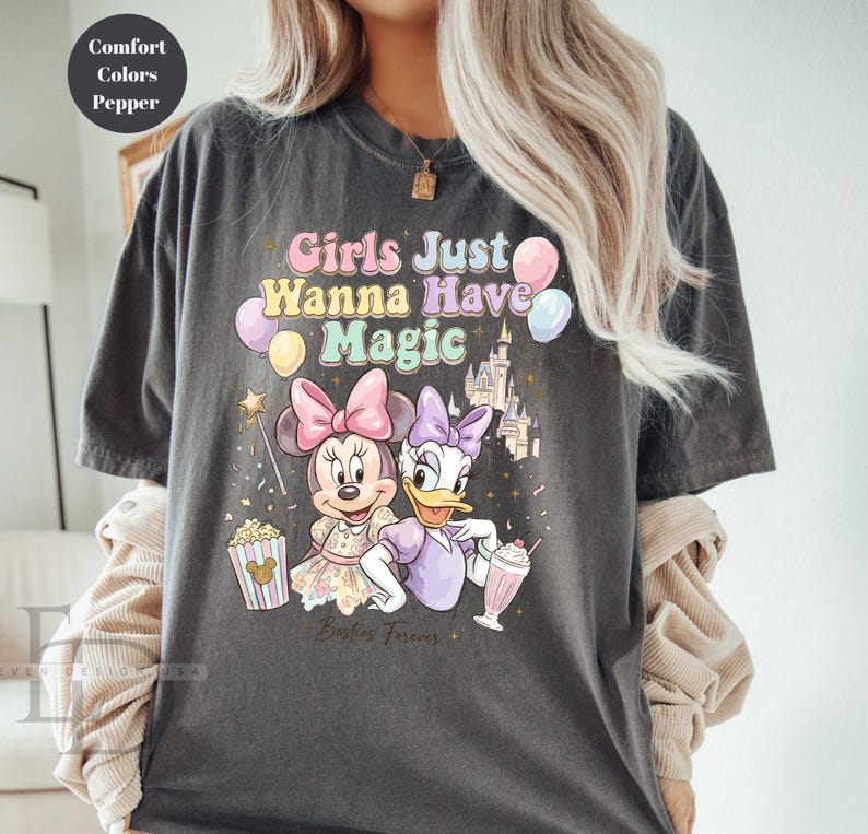 Puede incluir: Camiseta gris oscuro con Minnie Mouse y Daisy Duck y el texto "Girls Just Wanna Have Magic". El dise&ntilde;o incluye globos, un castillo y palomitas de ma&iacute;z. Las palabras "Besties Forever" tambi&eacute;n est&aacute;n impresas en la camiseta.
