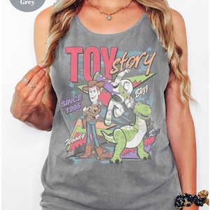 Puede incluir: Camiseta sin mangas gris con un gr&aacute;fico de los personajes de Toy Story: Woody, Buzz Lightyear y Rex. El texto "TOY story" es rosa y rojo, con "SINCE 1995" debajo. La camiseta es de color c&oacute;modo.