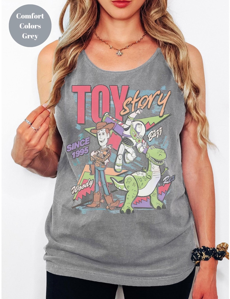 Puede incluir: Camiseta sin mangas gris con un gr&aacute;fico de los personajes de Toy Story: Woody, Buzz Lightyear y Rex. El texto "TOY story" es rosa y rojo, con "SINCE 1995" debajo. La camiseta es de color c&oacute;modo.