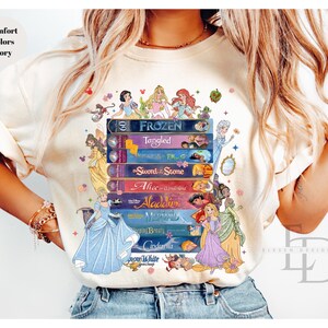 Puede incluir: Camiseta color marfil con un dise&ntilde;o de princesas Disney. El dise&ntilde;o presenta una pila de t&iacute;tulos de pel&iacute;culas como "Frozen" y "Tangled", con ilustraciones de princesas. Tambi&eacute;n se ve el texto "Comfort Colors Ivory".