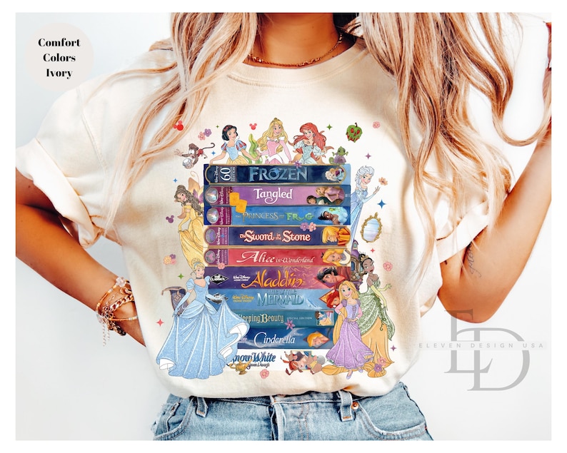 Puede incluir: Camiseta color marfil con un dise&ntilde;o de princesas Disney. El dise&ntilde;o presenta una pila de t&iacute;tulos de pel&iacute;culas como "Frozen" y "Tangled", con ilustraciones de princesas. Tambi&eacute;n se ve el texto "Comfort Colors Ivory".