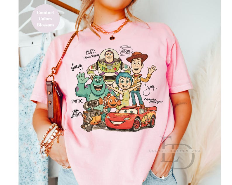 Puede incluir: Una camiseta de color rosa claro con un estampado gr&aacute;fico de personajes de Disney y Pixar, incluidos Buzz Lightyear, Woody, Sulley, Joy, Nemo, Wall-E y Lightning McQueen, con sus nombres escritos en una fuente manuscrita estilizada.