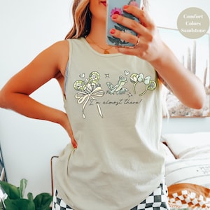 May include: Sandstone-colored tank top with a whimsical graphic featuring a frog, shamrocks, and a bow. The text "I'm almost there!" is written below the graphic. The top is made of a soft material.
