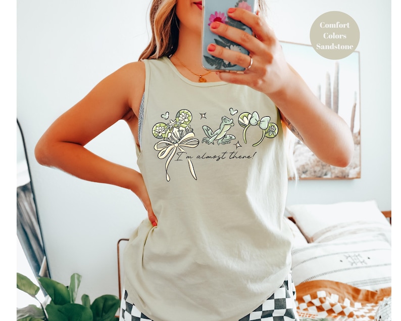 May include: Sandstone-colored tank top with a whimsical graphic featuring a frog, shamrocks, and a bow. The text "I'm almost there!" is written below the graphic. The top is made of a soft material.