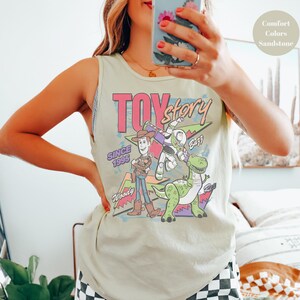 Puede incluir: Una camiseta sin mangas verde claro con un gr&aacute;fico colorido de los personajes de Toy Story. El dise&ntilde;o incluye a Woody, Buzz Lightyear y Rex, con las palabras "Toy Story" y "Since 1995". La camiseta sin mangas es de color Sandstone Comfort Colors.