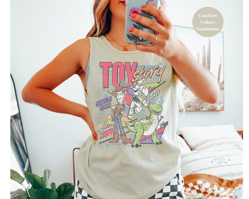 Puede incluir: Una camiseta sin mangas verde claro con un gr&aacute;fico colorido de los personajes de Toy Story. El dise&ntilde;o incluye a Woody, Buzz Lightyear y Rex, con las palabras "Toy Story" y "Since 1995". La camiseta sin mangas es de color Sandstone Comfort Colors.
