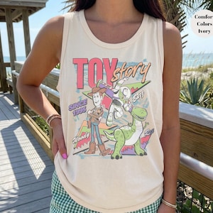 Puede incluir: Camiseta sin mangas color marfil con un gr&aacute;fico colorido de los personajes de Toy Story, incluyendo a Woody, Buzz Lightyear y Rex. Tambi&eacute;n se muestra el texto "Toy Story" y "Since 1995". La camiseta es de corte holgado.