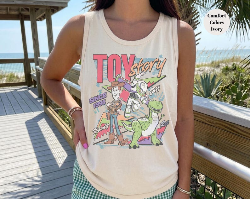 Puede incluir: Camiseta sin mangas color marfil con un gr&aacute;fico colorido de los personajes de Toy Story, incluyendo a Woody, Buzz Lightyear y Rex. Tambi&eacute;n se muestra el texto "Toy Story" y "Since 1995". La camiseta es de corte holgado.