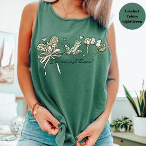 May include: A light green tank top with a whimsical design featuring a frog, flowers, and the text "I'm almost there!". The graphic is in shades of cream, yellow, and light green. The tank top is made of a soft, comfortable material.