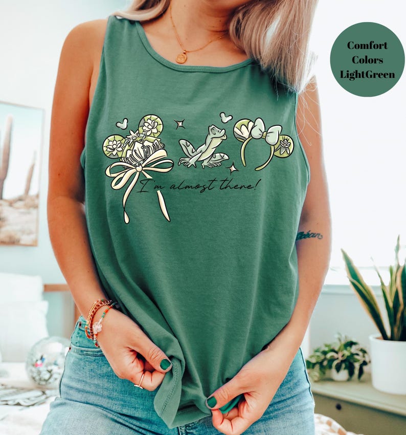 May include: A light green tank top with a whimsical design featuring a frog, flowers, and the text "I'm almost there!". The graphic is in shades of cream, yellow, and light green. The tank top is made of a soft, comfortable material.