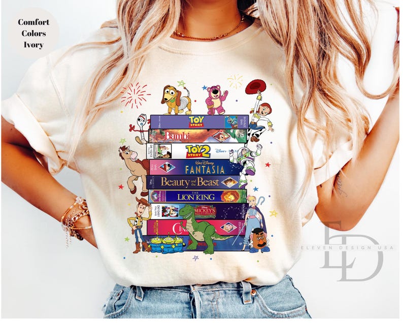 May include: Ivory-colored t-shirt featuring a stack of classic Disney VHS tapes with animated characters. The design includes characters from Toy Story, Bambi, Beauty and the Beast, and The Lion King. The shirt has the text "Comfort Colors Ivory".