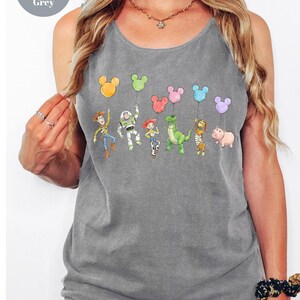 May include: Grey tank top featuring a colorful graphic of Toy Story characters holding Mickey Mouse-shaped balloons. The text "Comfort Colors Grey" is in a circle in the upper left corner. The tank top is sleeveless and has a scoop neck.
