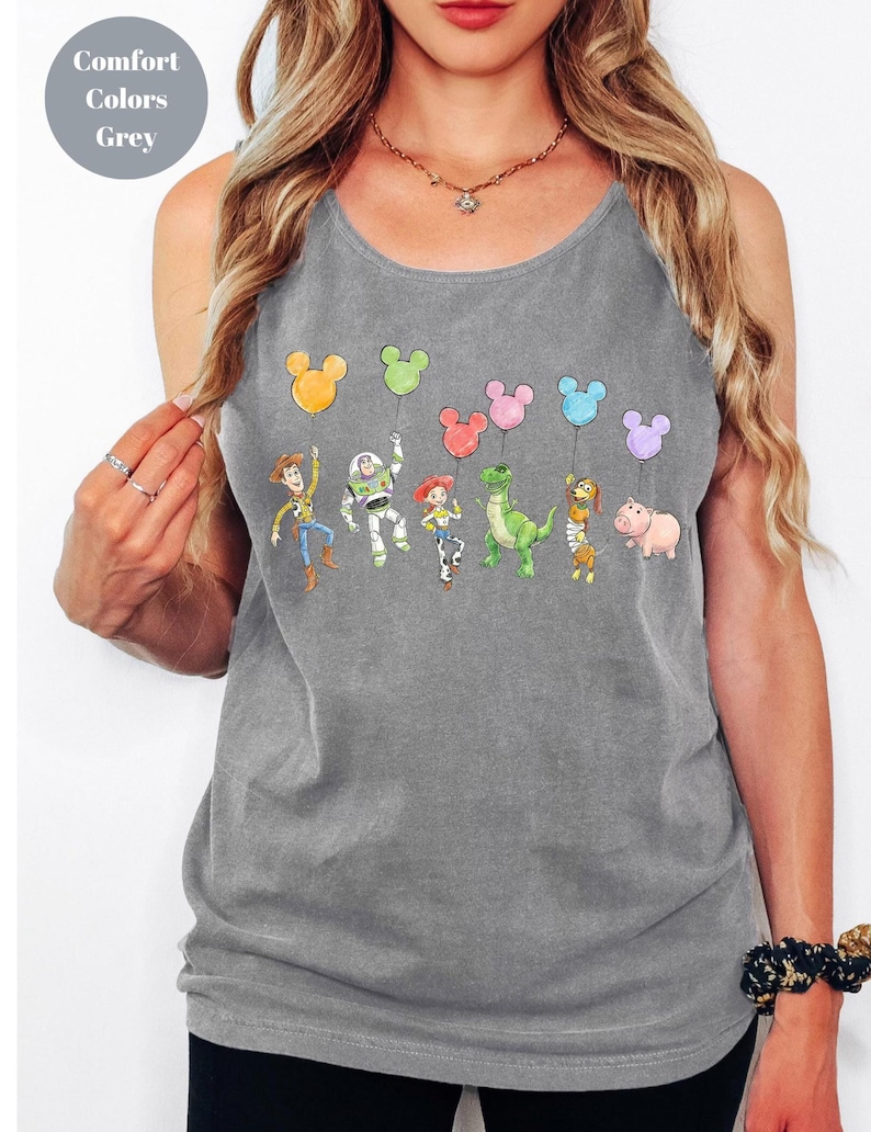May include: Grey tank top featuring a colorful graphic of Toy Story characters holding Mickey Mouse-shaped balloons. The text "Comfort Colors Grey" is in a circle in the upper left corner. The tank top is sleeveless and has a scoop neck.