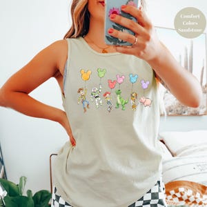 May include: A light beige tank top featuring a colorful graphic of cartoon characters holding Mickey Mouse-shaped balloons. The characters include Woody, Buzz Lightyear, and others. The text "Comfort Colors Sandstone" is in the upper right corner.