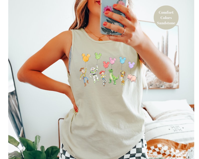 May include: A light beige tank top featuring a colorful graphic of cartoon characters holding Mickey Mouse-shaped balloons. The characters include Woody, Buzz Lightyear, and others. The text "Comfort Colors Sandstone" is in the upper right corner.