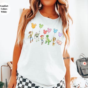 May include: White t-shirt featuring a colorful illustration of Toy Story characters holding Mickey Mouse-shaped balloons. The text "Comfort Colors White" is in the upper left corner. The design includes Woody, Buzz Lightyear, and other characters.
