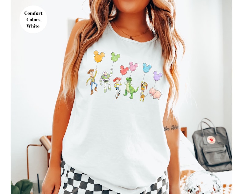 May include: White t-shirt featuring a colorful illustration of Toy Story characters holding Mickey Mouse-shaped balloons. The text "Comfort Colors White" is in the upper left corner. The design includes Woody, Buzz Lightyear, and other characters.