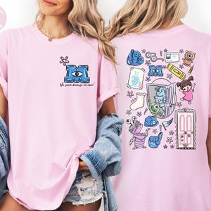 May include: Light pink t-shirt featuring a blue letter "M" with an eye design and the phrase "We scare because we care!" on the front. The back of the shirt has a colorful cartoon graphic with various characters and objects.