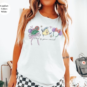 May include: White tank top featuring a colorful graphic design with the text "Part of your world." The design includes cartoon illustrations of a mermaid, a fish, and floral elements. The top is labeled "Comfort Colors White."