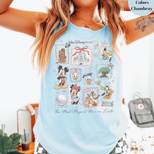 May include: Light blue tank top featuring a colorful graphic of Disney characters and landmarks, including Mickey Mouse, Minnie Mouse, and Cinderella's castle. The text "Walt Disney World" and "2026" are also visible. The shirt is made of chambray material.