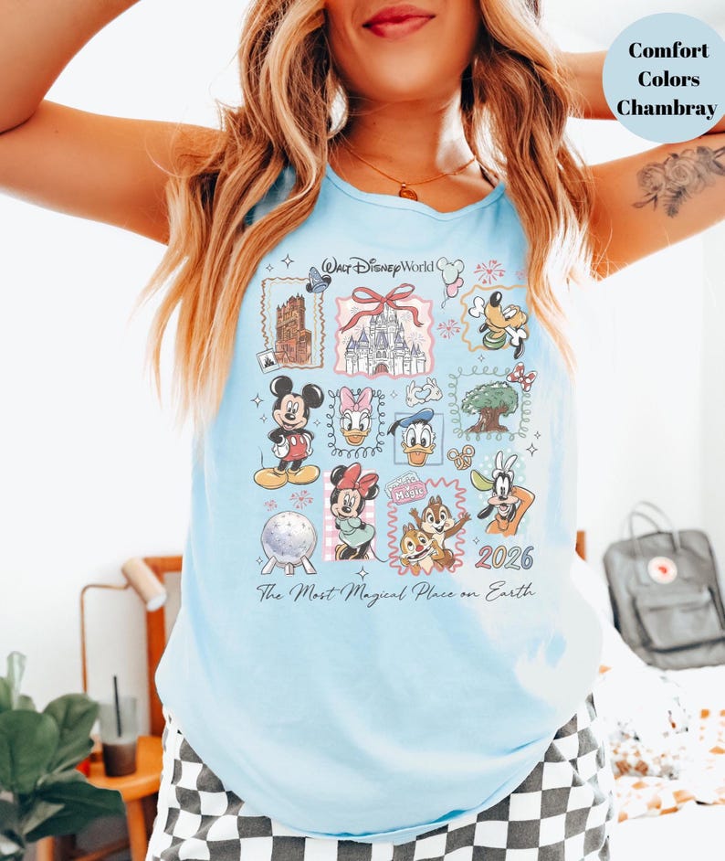 May include: Light blue tank top featuring a colorful graphic of Disney characters and landmarks, including Mickey Mouse, Minnie Mouse, and Cinderella's castle. The text "Walt Disney World" and "2026" are also visible. The shirt is made of chambray material.