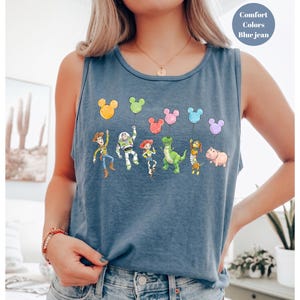 May include: Blue jean tank top featuring a colorful graphic of cartoon characters holding balloons. The characters include a cowboy, a space ranger, a cowgirl, a green dinosaur, and a pink pig. The top has the words "Comfort Colors" and "Blue jean" printed on it.