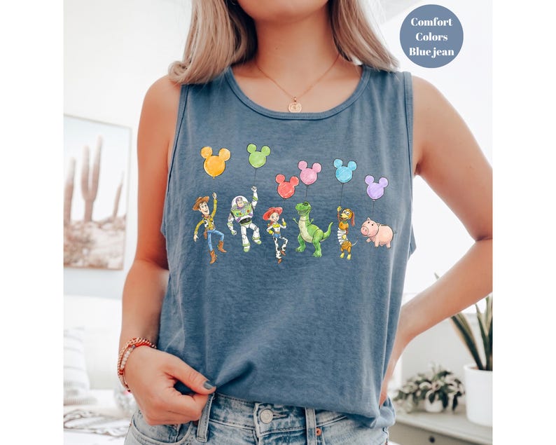 May include: Blue jean tank top featuring a colorful graphic of cartoon characters holding balloons. The characters include a cowboy, a space ranger, a cowgirl, a green dinosaur, and a pink pig. The top has the words "Comfort Colors" and "Blue jean" printed on it.