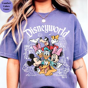 May include: A lilac t-shirt with the word "Disneyworld" in white script above a graphic of Disney characters including Mickey Mouse, Minnie Mouse, Donald Duck, and Goofy. The shirt also has the text "Florida 1971".