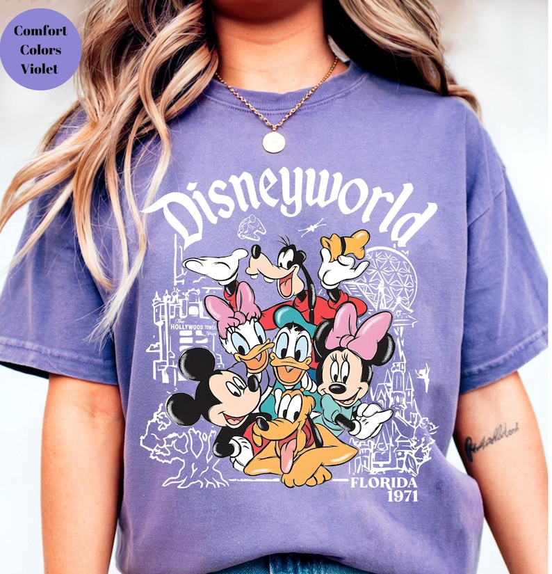 May include: A lilac t-shirt with the word "Disneyworld" in white script above a graphic of Disney characters including Mickey Mouse, Minnie Mouse, Donald Duck, and Goofy. The shirt also has the text "Florida 1971".