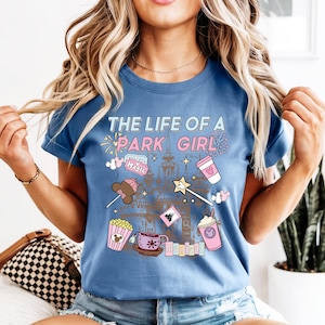 May include: A blue short-sleeved t-shirt with the text "THE LIFE OF A PARK GIRL" in a pastel font. The shirt features illustrations of a castle, popcorn, a coffee cup, a wand, and other park-themed graphics.