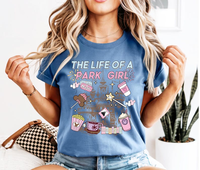 May include: A blue short-sleeved t-shirt with the text "THE LIFE OF A PARK GIRL" in a pastel font. The shirt features illustrations of a castle, popcorn, a coffee cup, a wand, and other park-themed graphics.