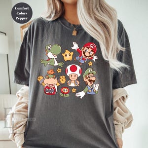 May include: Dark gray t-shirt featuring colorful cartoon characters from the Super Mario Bros. franchise. The design includes Mario, Luigi, Yoshi, Toad, Bowser, and a star. The shirt has the words "Comfort Colors Pepper" in a circle.