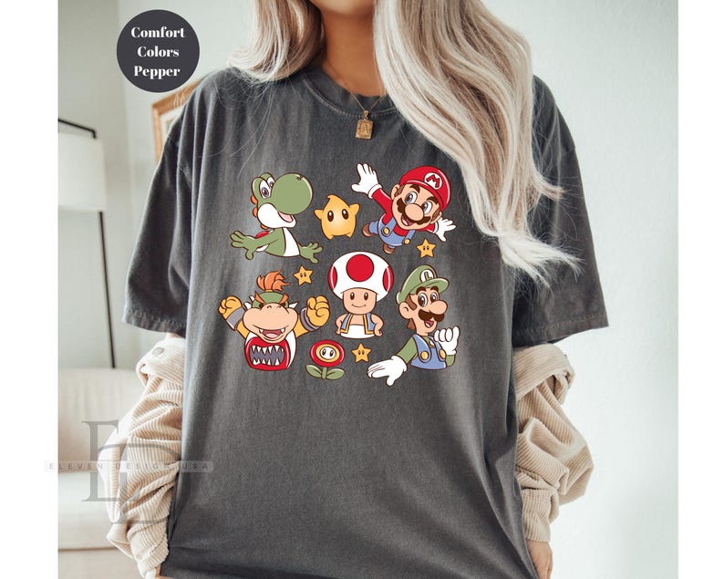 May include: Dark gray t-shirt featuring colorful cartoon characters from the Super Mario Bros. franchise. The design includes Mario, Luigi, Yoshi, Toad, Bowser, and a star. The shirt has the words "Comfort Colors Pepper" in a circle.