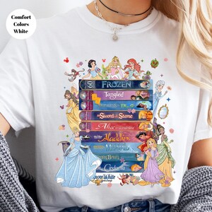 Puede incluir: Camiseta blanca con un dise&ntilde;o gr&aacute;fico colorido de libros de cuentos de Disney apilados. Los t&iacute;tulos incluyen "Frozen", "Tangled", "Alicia en el pa&iacute;s de las maravillas" y "Aladdin". El texto "Comfort Colors White" est&aacute; en un c&iacute;rculo blanco.