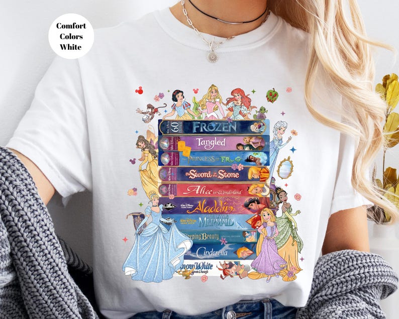 Puede incluir: Camiseta blanca con un dise&ntilde;o gr&aacute;fico colorido de libros de cuentos de Disney apilados. Los t&iacute;tulos incluyen "Frozen", "Tangled", "Alicia en el pa&iacute;s de las maravillas" y "Aladdin". El texto "Comfort Colors White" est&aacute; en un c&iacute;rculo blanco.