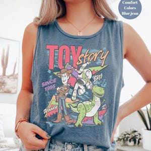 Puede incluir: Camiseta sin mangas azul vaquero con un gr&aacute;fico colorido de los personajes de Toy Story Woody, Buzz Lightyear y Rex. El texto "TOY STORY" es rosa, con "SINCE 1995" debajo. La camiseta sin mangas es de color confort.