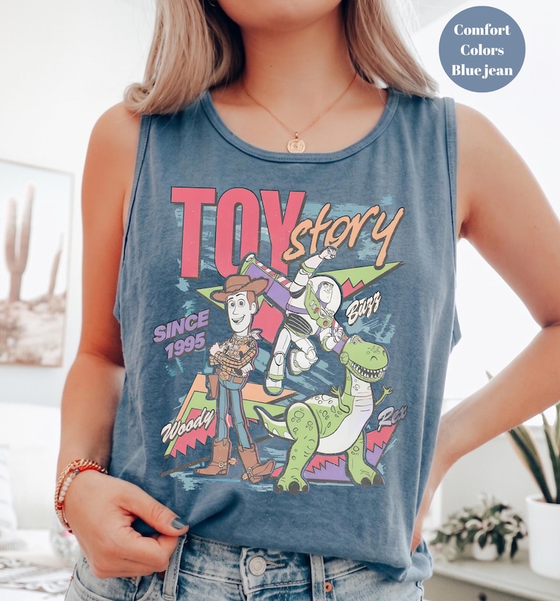 Puede incluir: Camiseta sin mangas azul vaquero con un gr&aacute;fico colorido de los personajes de Toy Story Woody, Buzz Lightyear y Rex. El texto "TOY STORY" es rosa, con "SINCE 1995" debajo. La camiseta sin mangas es de color confort.