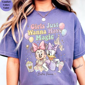 Puede incluir: Camiseta morada con Minnie Mouse y Daisy Duck, con el texto "Girls Just Wanna Have Magic" y "Besties Forever". El dise&ntilde;o incluye globos, un castillo y palomitas de ma&iacute;z.
