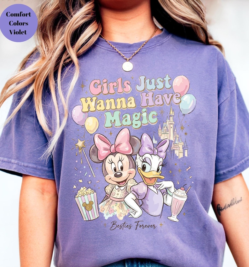 Puede incluir: Camiseta morada con Minnie Mouse y Daisy Duck, con el texto "Girls Just Wanna Have Magic" y "Besties Forever". El dise&ntilde;o incluye globos, un castillo y palomitas de ma&iacute;z.