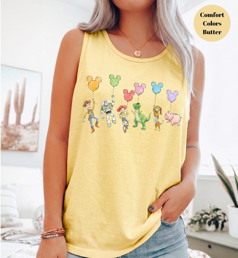May include: Yellow tank top featuring a whimsical illustration of cartoon characters holding Mickey Mouse-shaped balloons. The characters include a cowboy, a space ranger, a cowgirl, a green dinosaur, a dog, and a pig. The text "Comfort Colors Butter" is in the upper right corner.