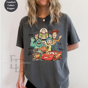 Puede incluir: Una camiseta de manga corta color gris carb&oacute;n con un estampado gr&aacute;fico de varios personajes animados, incluyendo a Buzz Lightyear, Woody, Sulley, Alegr&iacute;a, Nemo, Wall-E y Rayo McQueen, con los nombres de los personajes escritos en letra negra.