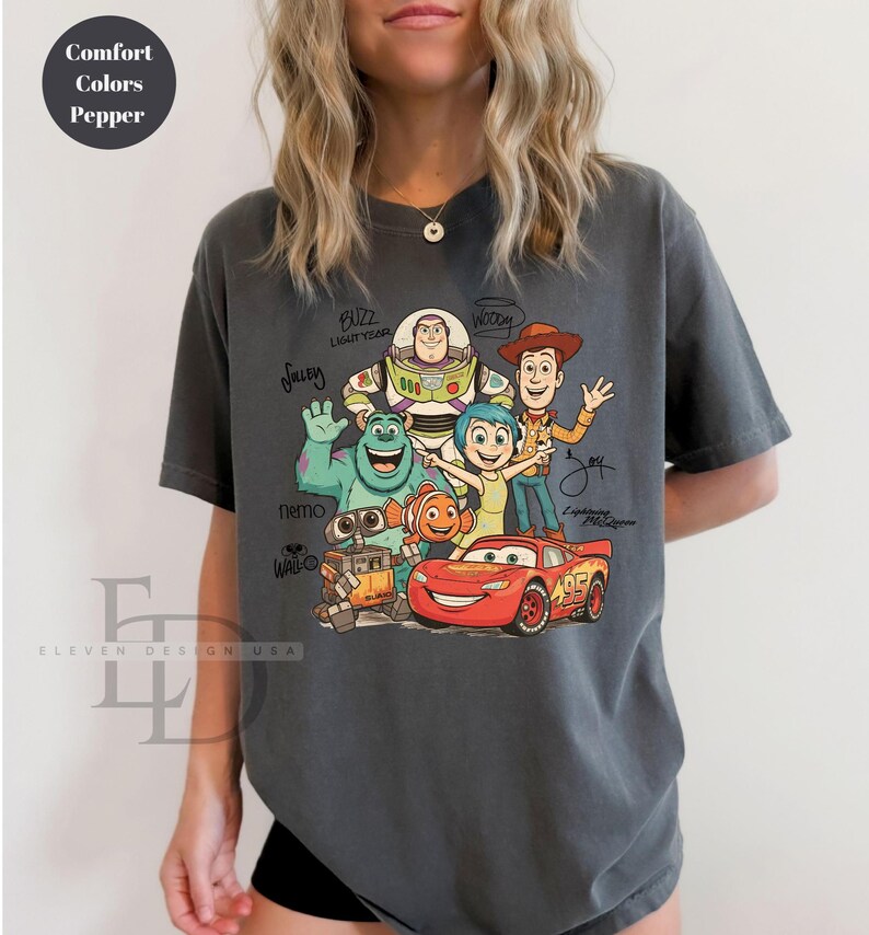 Puede incluir: Una camiseta de manga corta color gris carb&oacute;n con un estampado gr&aacute;fico de varios personajes animados, incluyendo a Buzz Lightyear, Woody, Sulley, Alegr&iacute;a, Nemo, Wall-E y Rayo McQueen, con los nombres de los personajes escritos en letra negra.