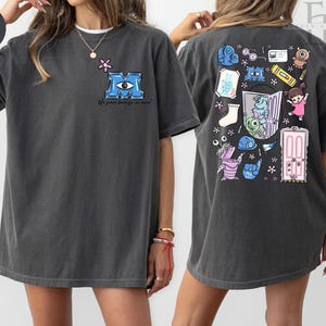 May include: Dark grey t-shirt featuring a blue "M" logo with an eye and the text "We scare because we care" on the front. The back of the shirt has a colorful cartoon design with various characters and objects.