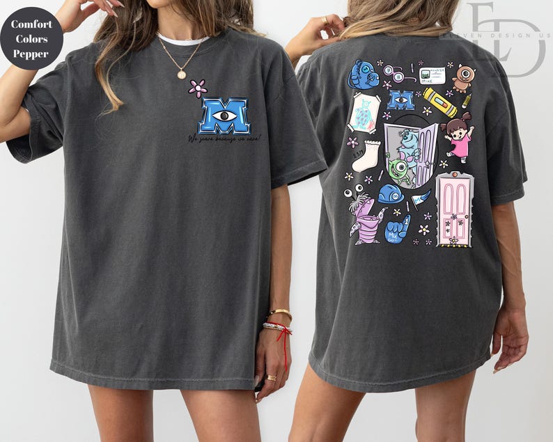 May include: Dark grey t-shirt featuring a blue "M" logo with an eye and the text "We scare because we care" on the front. The back of the shirt has a colorful cartoon design with various characters and objects.