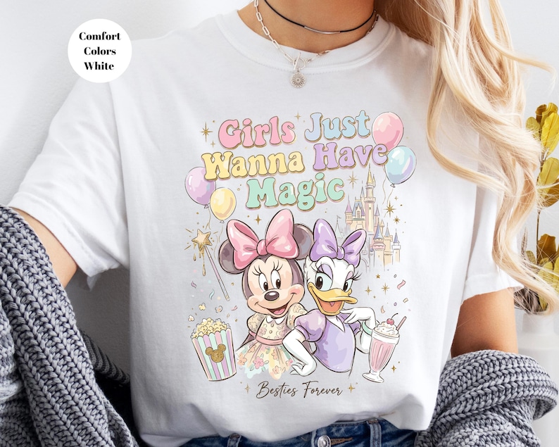 Puede incluir: Camiseta blanca con Minnie Mouse y Daisy Duck con el texto "Girls Just Wanna Have Magic". El dise&ntilde;o incluye un castillo, globos, palomitas de ma&iacute;z y un batido. Tambi&eacute;n est&aacute;n presentes las palabras "Besties Forever".