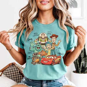 Puede incluir: Una camiseta de manga corta color verde azulado con un estampado gr&aacute;fico de varios personajes animados, incluyendo a Buzz Lightyear, Woody, Sulley, Joy, Nemo, Wall-E y Lightning McQueen, con sus nombres escritos en una fuente cursiva estilizada.