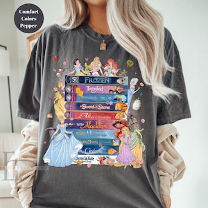 Puede incluir: Camiseta gris oscuro con un dise&ntilde;o de libros apilados con t&iacute;tulos de princesas de Disney como "Frozen" y "Cinderella." El dise&ntilde;o incluye ilustraciones de princesas y el texto "Comfort Colors Pepper."