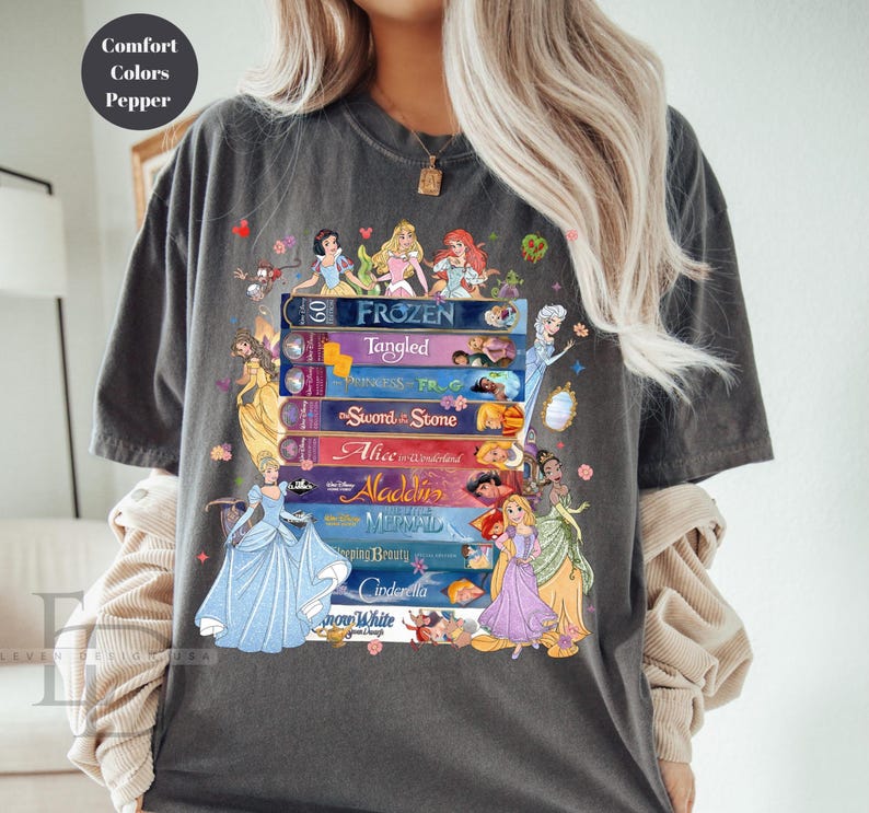 Puede incluir: Camiseta gris oscuro con un dise&ntilde;o de libros apilados con t&iacute;tulos de princesas de Disney como "Frozen" y "Cinderella." El dise&ntilde;o incluye ilustraciones de princesas y el texto "Comfort Colors Pepper."